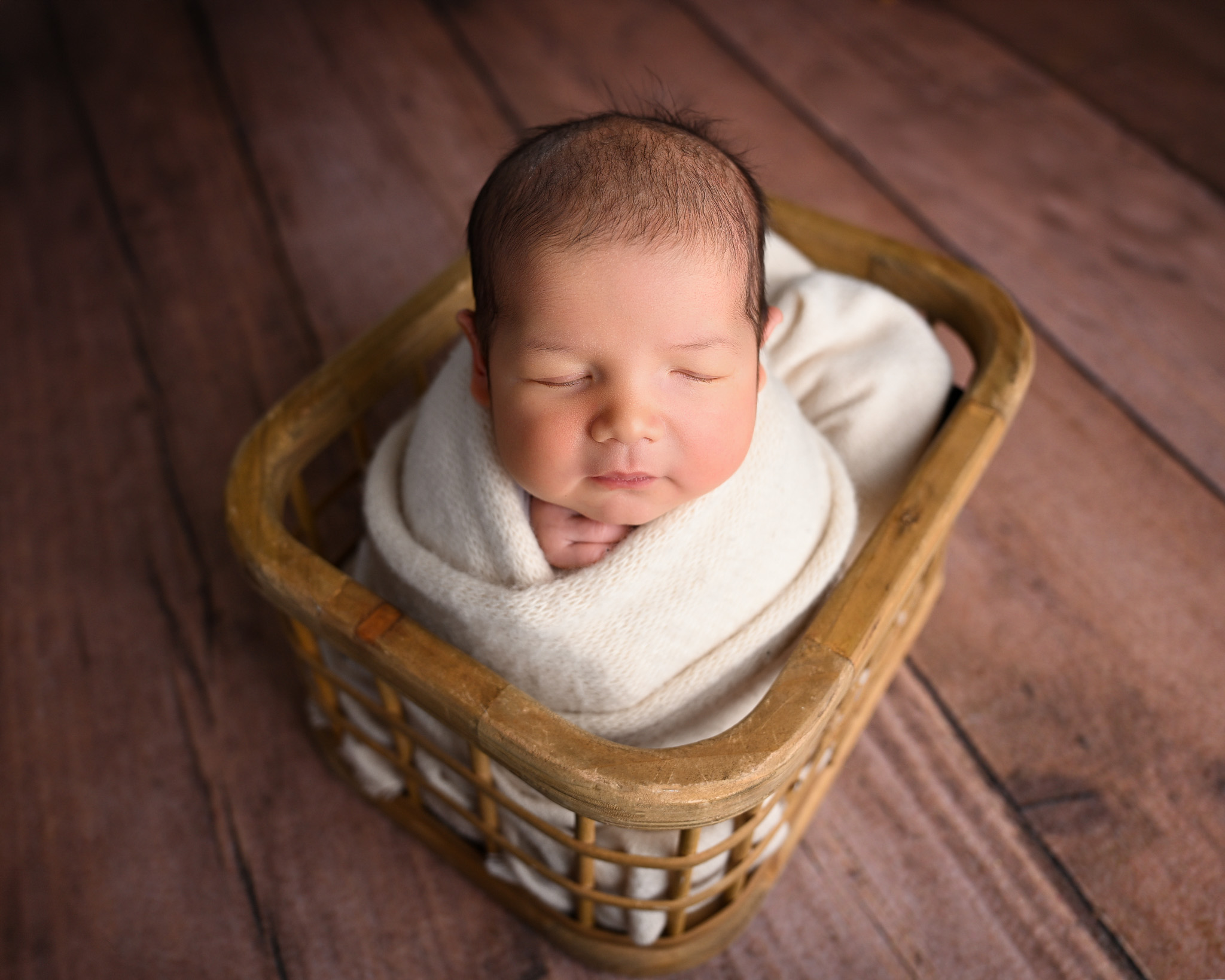 Newborn baby wrapped in a white blanket posed inside of a wood basket
